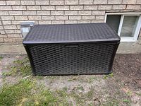 Front view of dark brown Suncast resin wicker deck box against brick wall