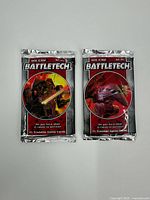 Front view of two sealed Battletech booster packs showing artwork, WOC 6302 code and 15-card notation.
