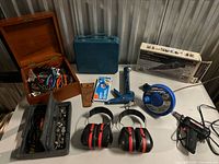Overall view of lot on table showing rotary tool kit box, wooden box of hand tools, blue carry case, glue gun, fish tape, rivet gun, ear muffs, organiser box