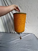 Full view of cylindrical swag lamp being held showing woven exterior and tassel