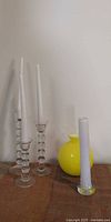 Three clear bubble-stem crystal candlesticks with white tapers beside yellow globe vase and tall white-lined bud vase