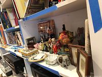 Overall view of shelf items including glass paperweight, pig figurine, trinket boxes, doll, plate, framed pictures