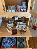 All items on two cabinet shelves: acrylic obelisks, octagonal cups, red mini tea set, white/gold tea service, bowls and spoons