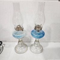Full view of both glass oil lamps showing chimneys, burners, blue reservoirs and bases