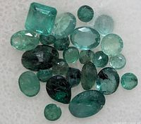 Close-up of approx 20 loose faceted emerald gemstones on white surface
