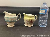 Front view of both Royal Albert creamers beside bottle for scale