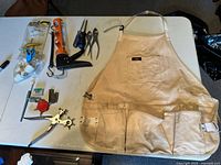 All items laid out on table including apron, caulking gun, pliers, scissors, bits, tape, valve tool