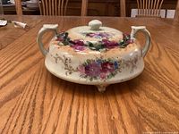 Full view of lidded porcelain pot showing two handles, floral design and three feet