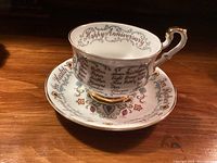 Teacup on saucer showing Happy Anniversary script, floral design, gold trim