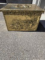 Front view of brass firewood box showing village scene and ornate border