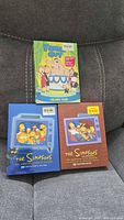 All three sealed DVD sets displayed on sofa: Family Guy Volume Four, Simpsons blue Collector’s Edition, Simpsons brown Collector’s Edition