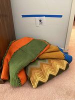 Three folded knit blankets stacked: orange-green stripe, earthtone chevron, blue-gold stripe