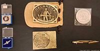 All six items laid out: Oklahoma buckle, Jerusalem buckle, two lapel pins in boxes, Draper Labs coin in sleeve, Swank tie clip