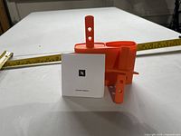 Front view of orange twin ice lolly mould, two stick handles, instruction booklet, tape measure for scale