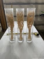 Three clear glass hurricane vases filled with dried wheat with measuring tape for scale