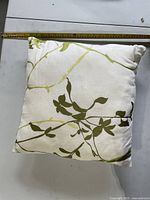 Front view of square white pillow with olive green vine pattern next to tape measure