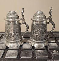 Front view of both pewter tankards showing relief scenes and lids