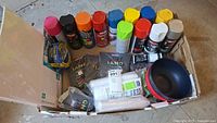 Box containing spray paint cans, art sand bags, painter’s tape, poster board, bowls and drop cloths