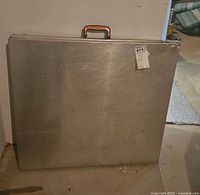 Table folded upright against wall showing speckled aluminum surface and plastic handle