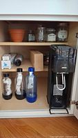 Cabinet interior showing SodaStream machine, bottles, bowls, jars, filter box, terracotta pot