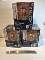 Three sealed Hampton Bay wall lantern boxes displayed with ruler for scale