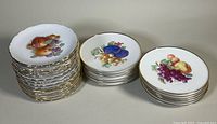 Stacked view of three groups of fruit motif dessert plates showing quantity and gold rim detail