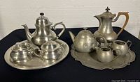 Both pewter serving sets on trays showing all nine pieces