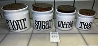 Front view of four ceramic canisters labelled Flour, Sugar, Coffee, Tea with cork lids