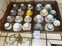 Two boxes holding numerous teacup and saucer sets, yellow creamer and pile of display stands