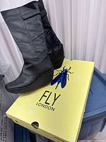 Side view of black leather Fly London tall wedge boot resting on branded yellow box