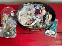 Open tin showing assorted bagged sewing notions such as thread spools, snaps and buttons