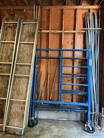 Stacked blue scaffold frames, plywood-aluminum planks leaning, long steel poles visible