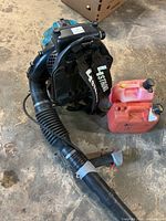 Makita backpack blower lying on floor with attached tube and two red gas cans