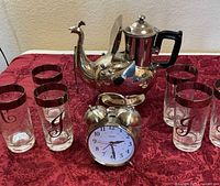 Full lot: peacock teapot, alarm clock, six J-monogram glasses on red cloth
