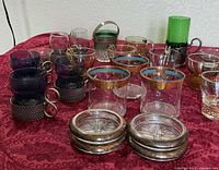 Group view showing metal holders with purple, cobalt, amber inserts, gold-rimmed glasses and stacked silver-plated coasters