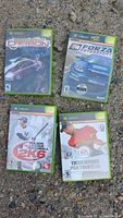 All four Xbox game cases on gravel: Need for Speed Carbon, Forza Motorsport, MLB 2K6, Tiger Woods 06