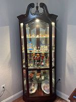 Full view of illuminated dark wood corner curio with glass panels and arched pediment