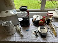 Entire lot displayed on table showing all ten items including blender, bread maker, ice-cream maker, immersion blender, popcorn popper with supplies, waffle maker, hand mixer, carving knife, scale, Eggies box
