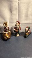 Three ornaments arranged side by side: gold bell with child, standing Santa, small Santa marked S'Nick