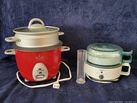 Rival rice cooker with lid and steamer basket beside Krups egg boiler, measuring beaker visible