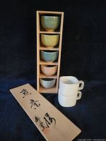 Full lot showing five pastel crackle-glaze teacups in wooden box, lid with calligraphy, and two white square cups