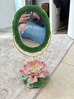 Full view of ceramic floral vanity mirror showing green framed oval mirror, gold rod, pink flower base