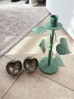 Green/teal painted metal candlestick with vine leaves beside two tarnished silver apple dishes on tile floor
