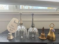 Five bells visible: pewter Gremlin Bell, two glass bells with silver-plated handles, copper-tone bell with wooden handle, brass bell with ring handle and purple flower