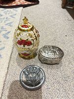 Three items: enamel egg holder, silver-tone trinket box closed, round metal trivet