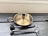 All three metal candle accessories laid out on dark surface: chamberstick, bell snuffer, wick trimmer scissors