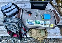 Group shot of tent bag, backpacks, cooler and accessories on bench