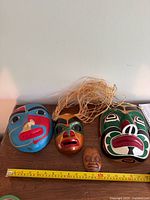 Front view of three large painted masks, one miniature mask and measuring tape