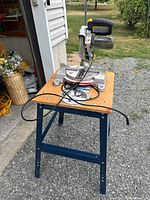 Front view of Mastercraft miter saw on stand with cord and packaged spare blade on table