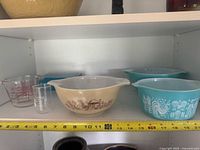 Shelf view showing beige farm scene bowl, two turquoise Butterprint bowls, turquoise refrigerator dish, Pyrex measuring cup and shot measure